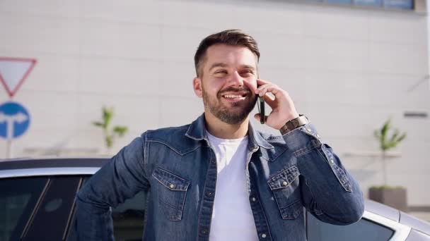 Vista frontale di simpatico giovane barbuto sorridente che parla al telefono mentre la sua classe lux electro car si collega alla stazione di ricarica — Video Stock