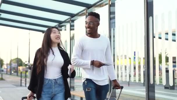 Привабливий радісний молодий бородатий афроамериканський і його азіатський подруга ходить на автобусному вокзалі з валізами на колесах перед польотом — стокове відео