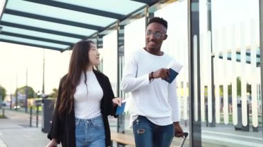 Çekici, genç, sakallı Afrikalı Amerikalı ve Asyalı bayan arkadaşı uçuştan önce tekerlekli bavullarıyla otobüs durağında yürüyorlar.