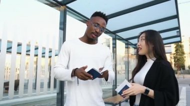 Yakışıklı, genç, sakallı Afrikalı Amerikalı Asyalı bayan arkadaşıyla otobüs durağında duruyor ve ortak tatil için biletlerini kontrol ediyor.