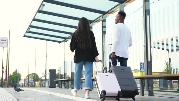 Vista desde atrás de dos jóvenes amigos multirraciales satisfechos que se reúnen en vacaciones y esperan el transporte en la parada de autobús — Vídeos de Stock