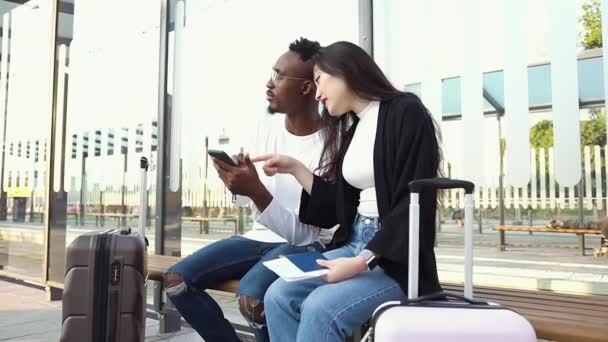 Atractivo joven barbudo moderno afroamericano sentado en la estación de tranvía junto con adorable asiática amiga y el uso de teléfono inteligente — Vídeos de Stock