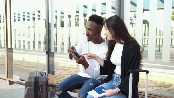 Widok z przodu atrakcyjnych pozytywnych zadowolonych mieszanych przyjaciół wyścigowych, którzy siedzą na przystanku tramwajowym lub autobusowym z walizkami i za pomocą smartfona do planowania wspólnych podróży — Wideo stockowe