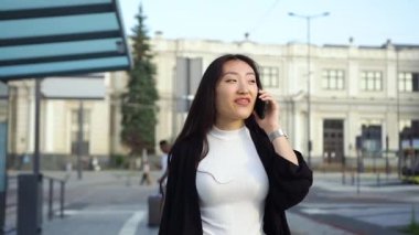 Sokakta yürüyen ve telefonda konuşan sıradan giysili mutlu Asyalı kadının portresi.