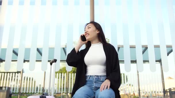 Vista frontal de la chica asiática sonriente bien parecido en ropa casual que habla por teléfono en la estación de transporte público — Vídeo de stock