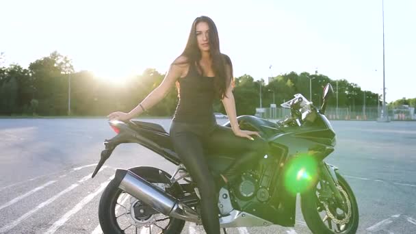 Великолепная улыбающаяся сексуальная молодая темноволосая женщина в черной одежде позирует перед камерой возле своего черного современного мотоцикла на дороге под солнечными лучами — стоковое видео