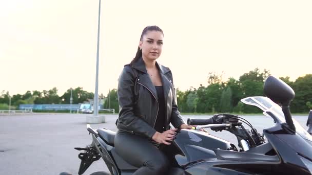 Довольно улыбающаяся молодая стильная брюнетка с хвостиком в модной одежде позирует на камеру на роскошном черном мотоцикле на спортивной площадке, крупным планом — стоковое видео