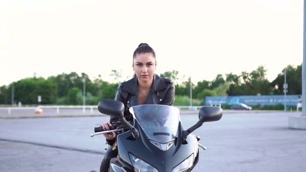 Splendide sourire heureux jeune femme mince avec queue de cheval en vêtements noirs posant sur la caméra sur moto moderne noire sur terrain de sport — Video