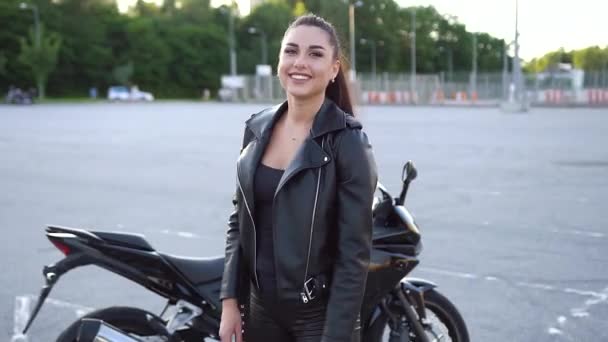 Dobře vypadající pozitivní elegantní mladá modish žena biker s culíkem v koženém oblečení pózuje v blízkosti luxusní černý motobike na sportovním hřišti — Stock video