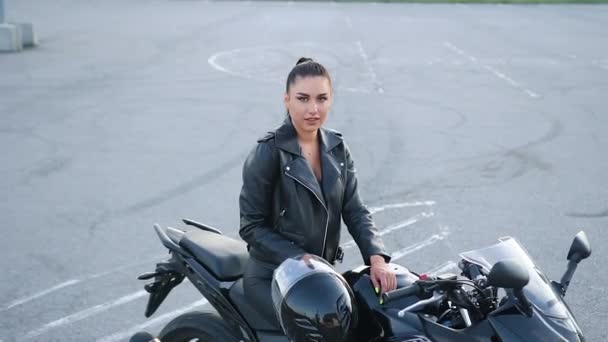 Belle jeune femme souriante modish insouciante en vêtements noirs assis sur une moto de luxe et regardant la caméra avec un visage heureux, gros plan — Video