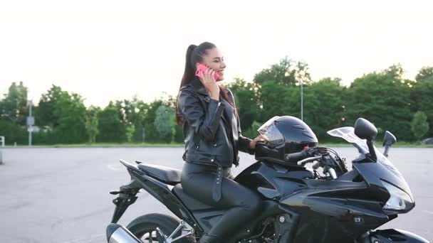 Gyönyörű gondtalan divatos fiatal női kerékpáros bőrruhában élvezi az okostelefon beszélgetést, miközben ül a drága fekete "vas ló" szabadban — Stock videók