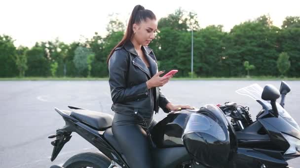 Красивая улыбающаяся стильная молодая женщина в черной одежде байкера сидит на привлекательном черном мотоцикле на спортивной площадке и пользуется телефоном — стоковое видео