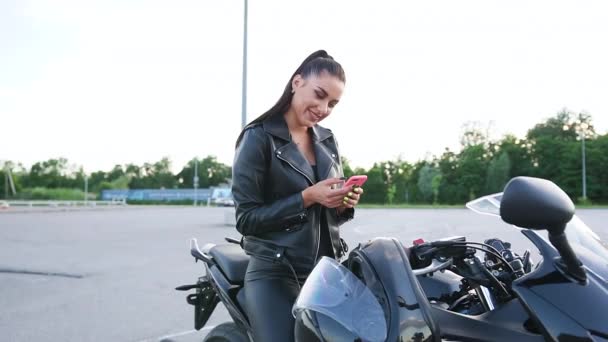 멋진 섹시 한 젊은 여성 자전거 선수가 가죽 옷을 입고 검은 운동복을 입고 모토 트랙 위에 앉아 스마트폰, 사이드 뷰를 사용하고 있습니다. — 비디오