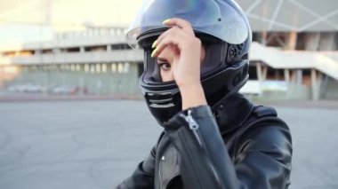 Güzel, kendine güvenen, modern, siyah kasklı genç bir kadın kameraya bakıyor, siyah spor bir motosikletin üzerinde oturuyor, yuvarlak manzaralı.