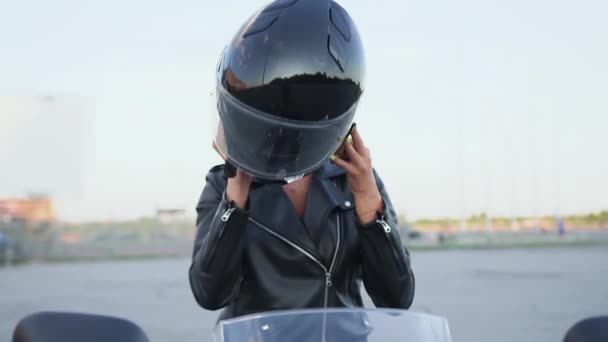 Attraktive selbstbewusste, stylische junge Bikerin, die sich einen schwarzen Helm aufsetzt, auf einem schwarzen Motorrad sitzt und in die Kamera schaut, Nahaufnahme — Stockvideo