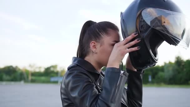 Vista lateral de atractiva mujer joven satisfecha con estilo con cola de caballo y ropa negra que se pone el casco protector, sentado en una lujosa moto negra — Vídeo de stock