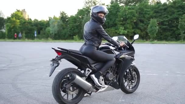 Attrayant confiant élégant jeune motocycliste mince femme en casque noir en vêtements noirs assis sur moto de luxe noir sur terrain de sport et en regardant la caméra — Video