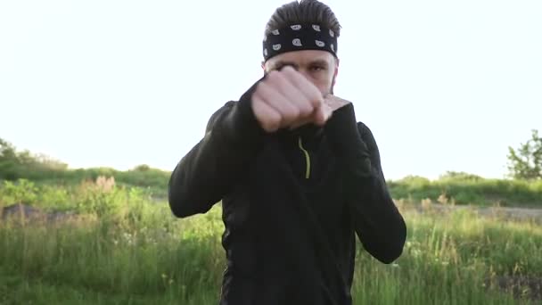 Hombre barbudo joven deportivo activo atractivo en ropa deportiva que practica golpes de boxeo en la cámara mientras entrena al aire libre, vista frontal — Vídeos de Stock