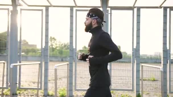 Вид збоку привабливий цілеспрямований активний сильний бородатий спортсмен в чорному одязі, який біжить біля спортивного стадіону, готуючись до марафонської дистанції — стокове відео