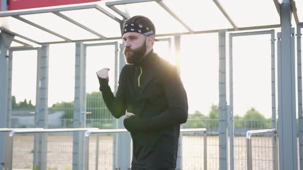 Крупним планом концентрований професійний молодий бородатий боксер в чорному одязі, який робить розтяжки і захисні удари на спортивному стадіоні — стокове відео