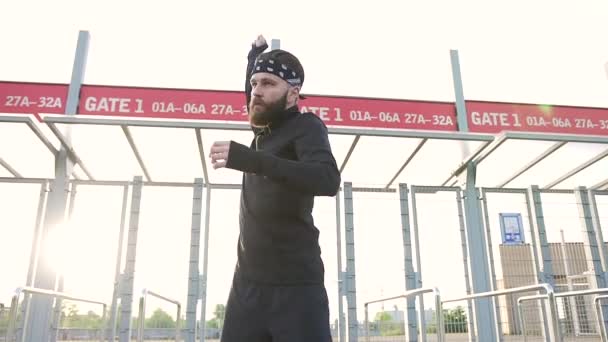 Aantrekkelijke geconcentreerde jonge sterke bebaarde man in sportkleding kneden spieren van zijn armen voor stadions toegangspoorten, close-up — Stockvideo