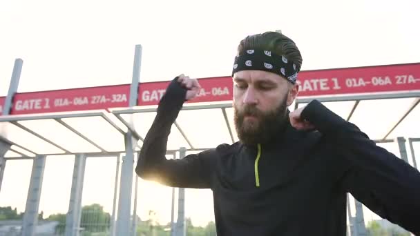 Vooraanzicht van aantrekkelijke geconcentreerde actieve jonge bebaarde atleet die op zijn plaats springt en zijn armen op het sportstadion kneedt — Stockvideo