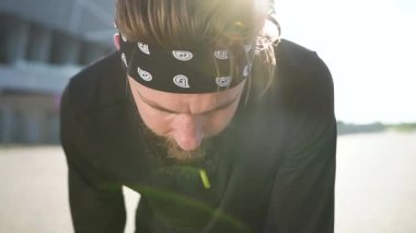 Spor giyimli, çekici, güçlü, sakallı, terini silen ve dışarıda egzersiz yaptıktan sonra nefes almaya devam eden genç adama yakın durun.