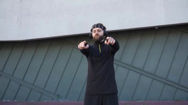 Çekici, ciddi, sakallı, kulaklıklı, siyah spor kıyafetleri giymiş, kollarını açıp kameraya poz veren ve sonra da çekip giden genç bir adam.