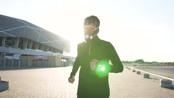 Närbild av säker målmedveten sportig skäggig löpare i skyddsmask klädd i svarta sportkläder som löper på den moderna arenan under solstrålar — Stockvideo