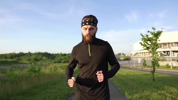 Portrét pohledný sebevědomý aktivní mladý vousatý sportovec v černém sportovním oblečení, které běží na stezce v blízkosti zelené louky na velkém stadionu pozadí — Stock video