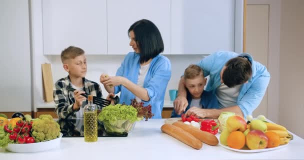 Çekici tatmin olmuş, tasasız iki kardeş, modern mutfaktaki gülümseyen genç aileleriyle sebze salatası pişirmeye yardım ediyor. — Stok video