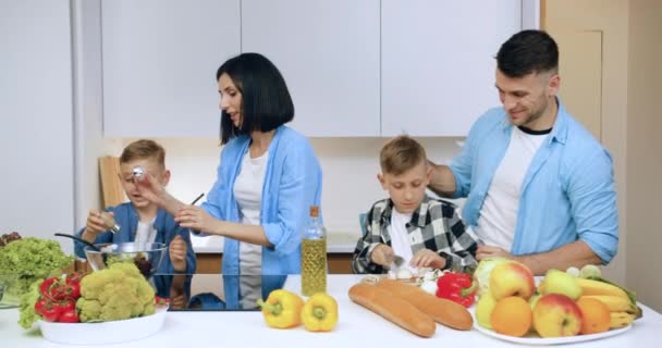 Ελκυστική χαρούμενη ευτυχισμένη γονείς μαγείρεμα σαλάτα λαχανικών μαζί με τα παιδιά από κοινού στη σύγχρονη κουζίνα, θέα μπροστά — Αρχείο Βίντεο