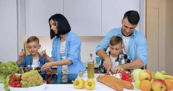 Retrato de una familia alegre y agradable con niños que se divierten juntos mientras cocinan útil ensalada de verduras naturales en la cocina moderna — Vídeos de Stock