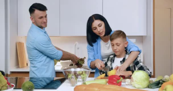 Attraente felice amichevole madre taglio pepe insieme con il suo piccolo figlio, mentre il suo bel marito lavaggio lattuga per la cena in cucina moderna — Video Stock