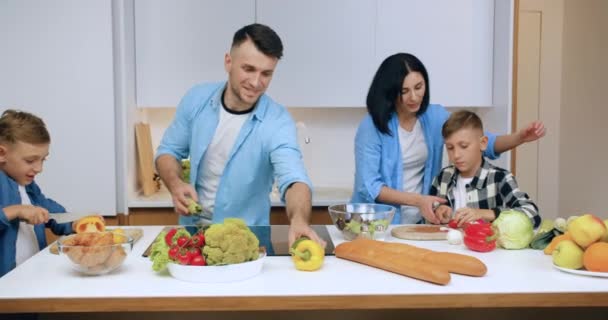 Idílio familiar onde atraente sorrindo pais felizes, juntamente com seus filhos cozinhar salada de legumes à mesa na cozinha contemporânea, close up — Vídeo de Stock