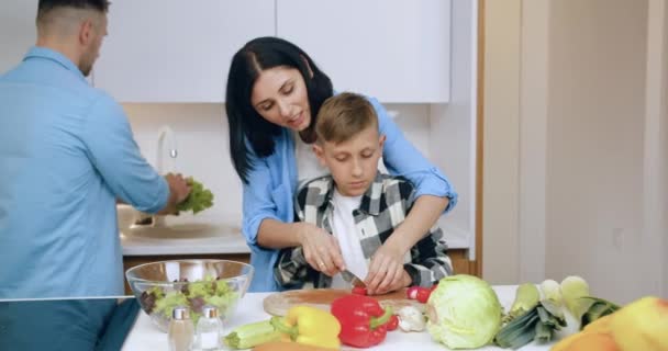 Bella sorridente felice mamma dai capelli scuri che insegna a suo figlio di 12 anni a tagliare il pomodoro mentre suo marito lava la lattuga per la cena congiunta nella cucina moderna — Video Stock