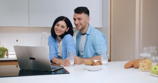 Preciosa sonrisa feliz pareja casada moderna sentada cerca de la mesa de la cocina y tener videollamada con un amigo o pariente en el ordenador portátil, de cerca — Vídeos de Stock