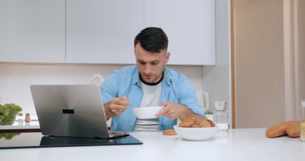 Гарний впевнений серйозний молодий чоловік, сніданок у затишній кухні одночасно працює на комп'ютері, вид спереду — стокове відео
