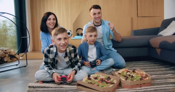 Schön lächelnde glückliche Eltern, die ihre beiden Söhne ermutigen, Videospiel zu spielen und nach Spielende Pizza zu genießen — Stockvideo