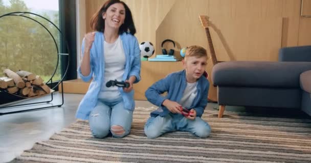 Close up of attractive positive happy cheerful mother and son which sitting on the floor and having fun during playing video game using joysticks — Stock Video