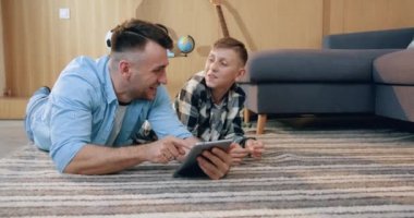 Oturma odasında yerde yatan ve i-pad 'i kullanarak ilginç uygulamaları izleyen yakışıklı, mutlu baba ve oğluna yakın durun.