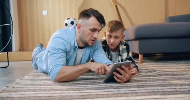 Atractivo papá moderno y seguro de sí mismo enseñando a su hijo adolescente cómo usar i-pad mientras pasan su tiempo libre en casa, acostados en la alfombra — Vídeos de Stock