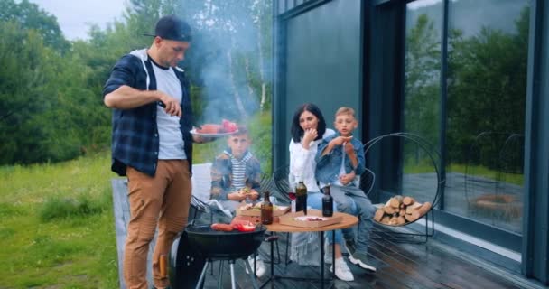 Aantrekkelijke zorgeloze vader zet op de grill worstjes en groenten, koken bbq partij voor zijn familie die zitten rond de tafel — Stockvideo