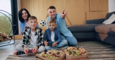 Çocuklar video oyunu oynarken evde oturup eğlenen neşeli ve çekici bir ailenin ön görüntüsü.