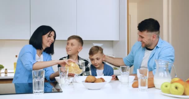 Οικογενειακή έννοια όπου όμορφη χαμογελαστή μελαχρινή μητέρα βάζοντας σε μπολ δημητριακά κορν φλέικς για τους δύο γιους της, ενώ έχοντας πρωινό στη σύγχρονη κουζίνα — Αρχείο Βίντεο