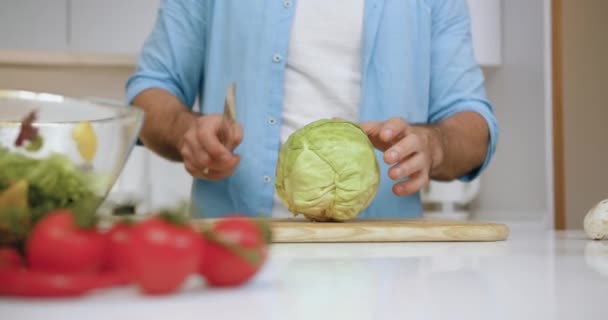 Vedere frontală a mâinii bine întreținute de sex masculin care taie varza verde pe două părți folosind cuțitul pe placa de tăiere în bucătăria modernă — Videoclip de stoc