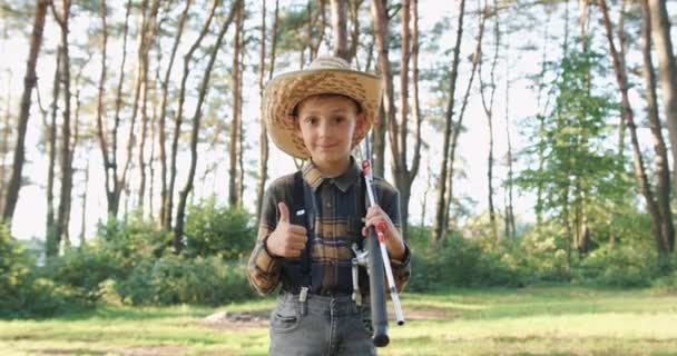 Vue de face de bon caractère satisfait sourire heureux garçon de 10 ans en chapeau de paille qui pose sur la caméra avec des cannes à pêche dans le parc vert et montrant le signe ok — Video