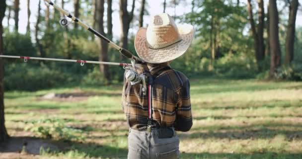 Vue arrière de attrayant petit garçon inconnu en chapeau de paille qui tient cannes à pêche sur ses épaules quand va pêcher parmi les arbres verts — Video