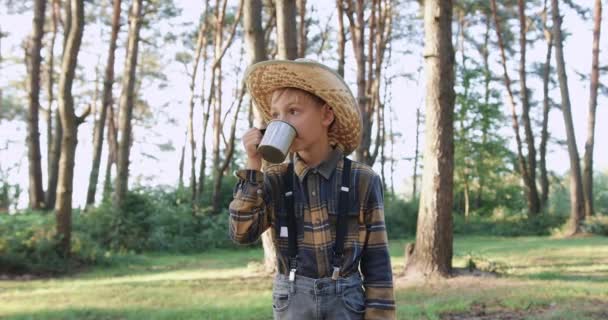 Gros plan de beau petit garçon réfléchi dans le chapeau qui boit du thé parmi le parc vert le jour ensoleillé — Video