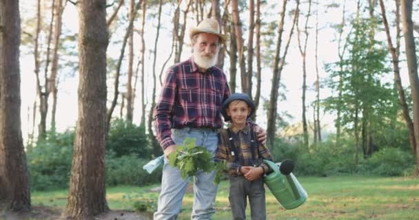 Portret van goed uitziende lachende vriendelijke bebaarde grootvader en kleinzoon die op zoek naar camera tijdens gezamenlijk werk in het groene park waar het planten van zaailing van eik — Stockvideo